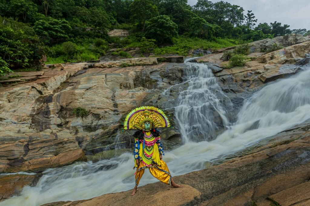 Chhau dancer in vibrant costume performing by a waterfall in Purulia, India.