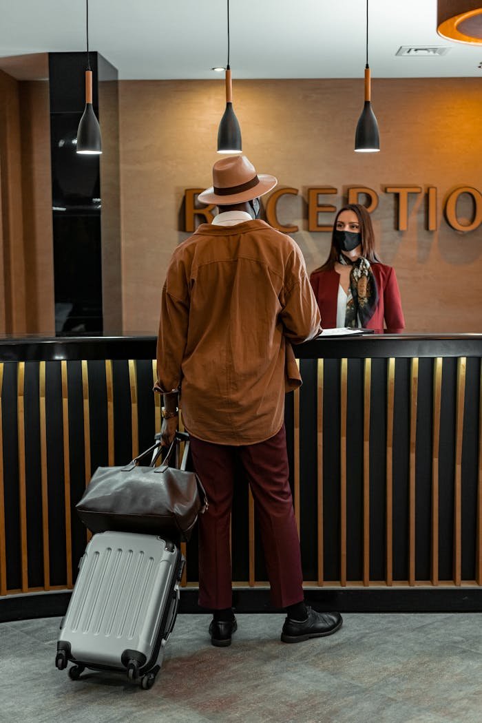 about-03 Man with luggage checking in at hotel reception. Staff wearing mask, indoors, travel theme.